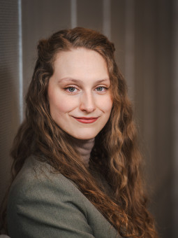 Junge Frau mit langen, lockigen Haaren in grauem Blazer, stehend an strukturierter Wand, Blick zur Kamera.