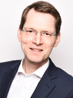 Mann mit kurzen braunen Haaren und Brille, in dunkelblauem Anzug, lächelt in die Kamera. Heller Hintergrund.