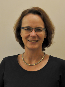 Frau mit schulterlangen, lockigen braunen Haaren, schwarzer Bluse und bunter Halskette lächelt in die Kamera.