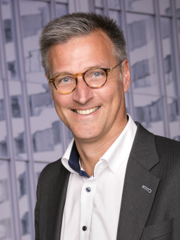 Mann mit grauen Haaren, Brille und Anzug lächelt vor moderner Architektur.