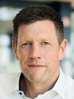 Mann mit kurzen, dunklen Haaren und Bart in weißem Hemd, schaut neutral in die Kamera, unscharfer Hintergrund.