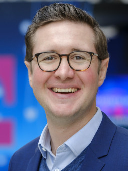 Mann mit Brille in blauem Anzug und hellblauem Hemd lächelt vor pinkem "T"-Logo in moderner Umgebung.