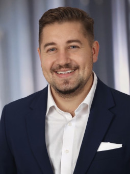 Mann mit braunen Haaren und Bart in dunkelblauem Anzug lächelt in die Kamera, unscharfer Hintergrund.