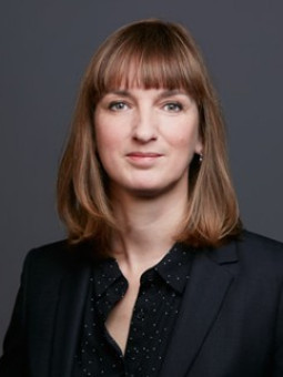 Frau mit schulterlangen, braunen Haaren in schwarzem Blazer und gepunktetem Hemd, neutraler Gesichtsausdruck.