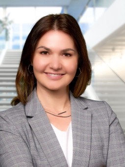 Frau mit langen, braunen Haaren in grauem Blazer, lächelt vor heller Umgebung mit Treppe und großen Fenstern.