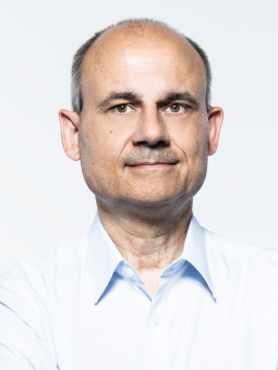 Mann mit kurzen braunen Haaren und Bart, trägt hellblaues Hemd, Arme vor der Brust verschränkt, neutraler Gesichtsausdruck.