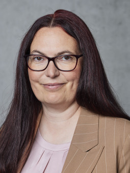 Frau mit langen, gewellten, dunklen Haaren, Brille, beigem Blazer und hellem Oberteil vor grauer Wand.