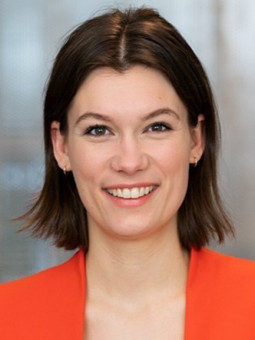 Person mit schulterlangen, braunen Haaren in rotem Blazer, lächelt in einem hellen, unscharfen Raum.