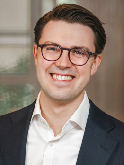 Person mit kurzen, dunklen Haaren und Brille, in hellem Hemd und dunklem Blazer, lächelt vor unscharfem Hintergrund.