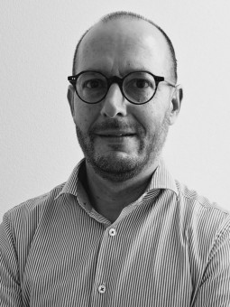 Person mit gestreiftem Hemd und runder Brille steht vor einer hellen Wand, Arme vor der Brust verschränkt.
