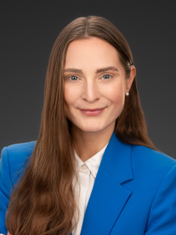 Person mit langen, braunen Haaren in blauem Blazer und weißem Hemd, vor dunklem Hintergrund, lächelt leicht.