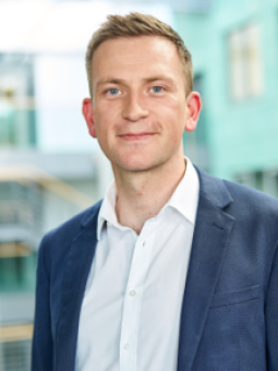 Mann mit kurzen, hellen Haaren in weißem Hemd und dunkelblauem Blazer, lächelt in modernem Raum.