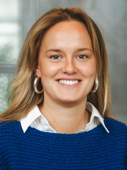Person mit langen, hellen Haaren, blauem Pullover, weißem Hemd und runden Ohrringen, lächelt vor unscharfem Hintergrund.