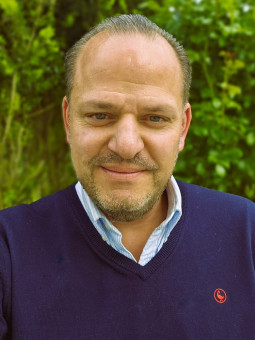 Person mit dunkelblauem Pullover und hellblauem Hemdkragen lächelt vor grünen Pflanzen.