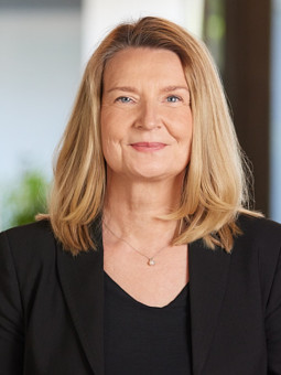 Frau mit schulterlangen blonden Haaren in schwarzem Blazer, lächelt vor unscharfem, hellem Hintergrund.