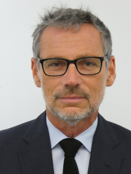 Mann mit grauen Haaren und Bart, schwarzer Brille, dunklem Anzug, hellblauem Hemd, schwarzer Krawatte, schaut in die Kamera.