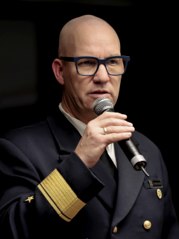 Mann in Uniform mit goldenen Schulterstücken spricht ins Mikrofon, trägt Brille mit blauem Gestell. Dunkler, unscharfer Hintergrund.