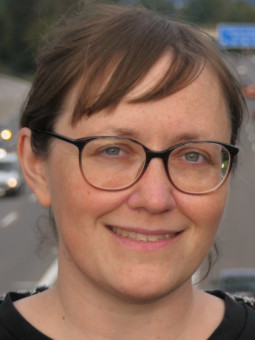Frau mit braunen, gewellten Haaren in schwarzer Bluse und gemustertem Kragen vor einer mehrspurigen Autobahn.
