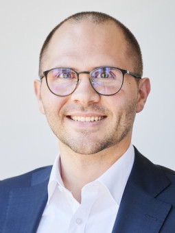 Person mit Brille lächelt in die Kamera, trägt dunklen Anzug und weißes Hemd, neutraler Hintergrund.