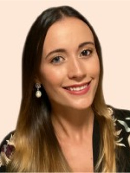 Person mit langen, glatten Haaren, schwarzer Bluse mit floralem Muster, lächelnd, große runde Ohrringe, heller Hintergrund.