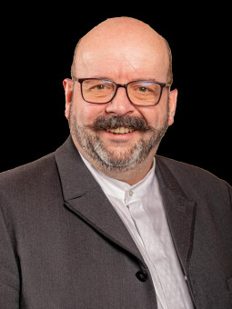 Markus Wartha, glatzköpfig mit Vollbart, trägt eine graue Jacke über einem weißen Hemd vor schwarzem Hintergrund.