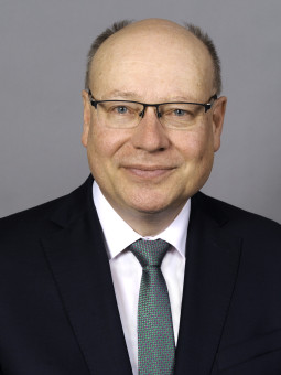 Frank Wetzel lächelt in die Kamera, trägt einen dunklen Anzug und eine gemusterte Krawatte. Grauer Hintergrund.