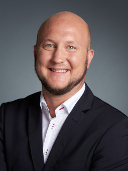Benedikt Frey in dunklem Anzug mit hellem Hemd, lächelnd, Arme vor dem Körper verschränkt, grauer Hintergrund.