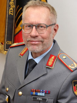 Person in grauer Uniform mit roten und goldenen Akzenten, Rangabzeichen, Brille, Bart, Medaillen, Namensschild "STINGL".