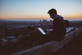 Junger Mann sitzt mit Laptop über den Dächern einer Stadt