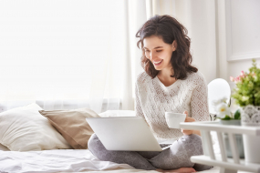 Frau sitzt mit Laptop und einer Tasse Kaffee im Bett