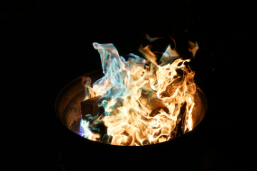 In der Mitte des Bildes lodert ein Feuer in einer dunklen Umgebung. Die Flammen sind teils orange, teils leuchtend blau-grün, was auf eine besondere chemische Reaktion oder Brennstoffe hinweist. Holzscheite liegen im Feuer und werden von den Flammen umschlossen. Das Feuer befindet sich in einem runden Metallbehälter, der nur teilweise im Bild zu sehen ist. Die Umgebung ist ansonsten dunkel und es gibt keine weiteren sichtbaren Objekte.