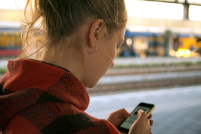 Frau prüft etwas auf dem Smartphone und steht dabei an einem Bahnsteig. 