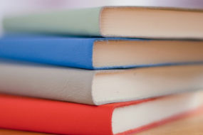 Vier geschlossene Notizbücher liegen horizontal gestapelt auf einem hellen Holztisch. Die Einbände sind glatt und unifarben, von oben nach unten in Hellgrün, Blau, Grau und Rot. Die Bücher sind gleich groß und gleichmäßig übereinandergelegt, mit den Schnittkanten der Seiten nach vorn gerichtet. Der Hintergrund ist unscharf in hellen Rosatönen gehalten, ohne erkennbare Details. Keine Personen im Bild.
