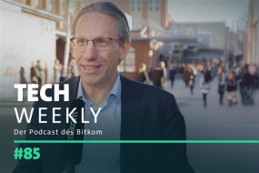 Bundesfinanzminister Dr. Jörg Kukies gibt ein Interview im Freien. Er trägt ein dunkles Sakko über einem hellblauen Hemd und hält ein schwarzes Mikrofon mit einem Logo darauf. Er lächelt freundlich und blickt in Richtung des Interviewers. Im Hintergrund gehen mehrere Menschen über einen Platz mit Backsteingebäuden. Das Bild ist mit einem dunklen Farbfilter überlagert, und darüber liegt der Text „TECH WEEKLY – Der Podcast des Bitkom“ sowie die Episodennummer „#85“ in grüner Schrift.