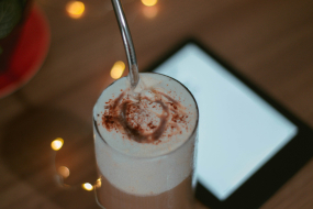 Ein Glas Latte mit Milchschaum steht im Vordergrund auf einem Holztisch. Im Hintergrund ein verschwommenes E-Book.