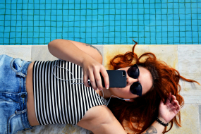 Eine junge Frau liegt am Rand eines Pools und hält ein schwarzes Smartphone direkt vor ihrem Gesicht.