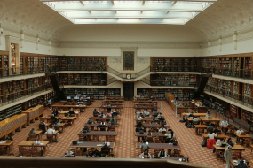 In einer großen, historischen Bibliothek mit hoher Decke sitzen zahlreiche Studierende an langen Holztischen. 