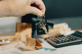 Hand hält Hausschlüssel über einem Tisch mit Geldscheinen, Taschenrechner und Finanzdokumenten.