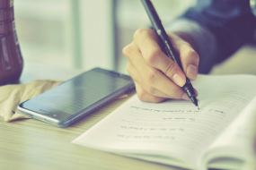 Smartphone auf einem Schultisch neben Schüler der in ein Heft schreibt