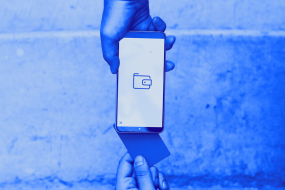 Jemand hält eine Karte in der Hand und legt diese an der Rückseite eines Handys an, das jemand anders in der Hand hält. Auf dem Handydisplay ist ein Wallet-Icon zu sehen. Das Bild zeigt einen digitalen, kontaktlosen Bezahlvorgang.
