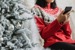 Person mit Weihnachtspullover sitzt neben einem Weihnachtsbaum und Geschenken und hält dabei ein Smartphone in der Hand.
