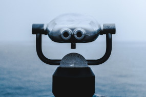 Silbernes Fernglas auf Standfuß mit schwarzem Haltegriff, unscharfer grauer Landschaft im Hintergrund. Text: „VIEWING MACHINE“.