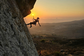 Frau klettert an einer Felswand, zwei Personen beobachten sie. Klettermatten liegen am Boden, Sonnenaufgang im Hintergrund.
