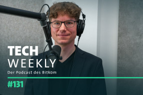 Mann mit lockigem, braunem Haar in schwarzem Sakko vor Mikrofon und Kopfhörern. Hintergrund in Grautönen.