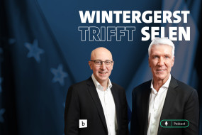 Zwei Männer in schwarzen Sakkos und weißen Hemden stehen nebeneinander. Hintergrund mit unscharfen Flaggen.