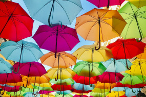 Bunte Regenschirme in Blau, Pink, Gelb, Grün und Orange hängen über einer Straße, umrahmt von Gebäudewänden.