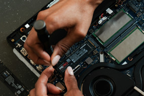 Zwei Hände arbeiten an einem geöffneten Laptop, eine hält einen Schraubendreher, die andere berührt die Hauptplatine.