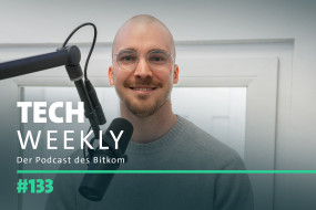 Mann mit Glatze und Brille lächelt vor Mikrofon, trägt grauen Pullover. Hintergrund: helle Wände und Fenster.