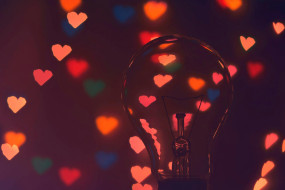 Transparente Glühbirne im Vordergrund, unscharfe, leuchtende Herzen in Rot, Pink, Blau und Gelb im Hintergrund.