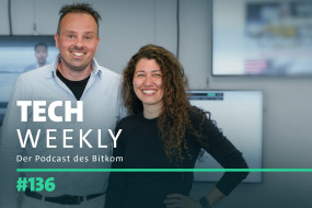 Zwei Personen lächeln vor Bildschirmen. Links: hellblaues Hemd, kurze Haare. Rechts: schwarzes Oberteil, lange lockige Haare.